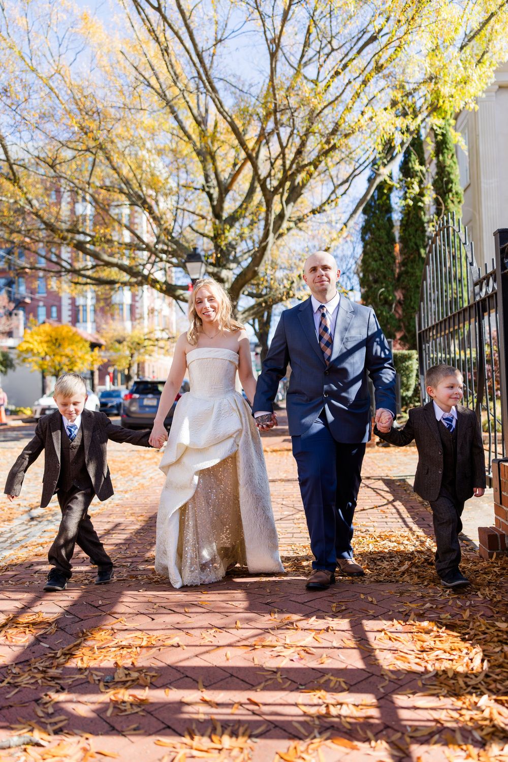 Wedding couple walking hand in hand with two young boys on a brick path surrounded by autumn foliage.