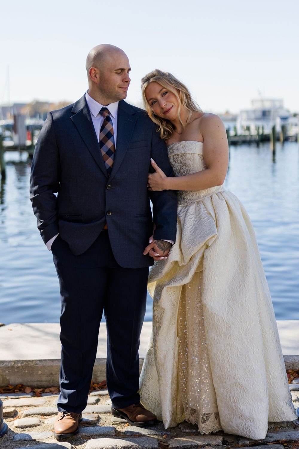 Bride in strapless ivory gown leans on groom in navy suit while posing at a sunny waterfront marina.
