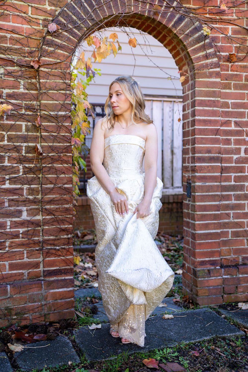 Bride in strapless ivory and gold gown poses gracefully beneath a rustic brick archway with autumn foliage.
