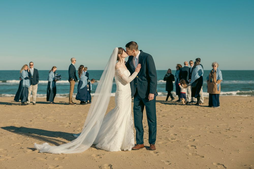 A bride and groom share a kiss on a sunny beach with their wedding party gathered behind them.