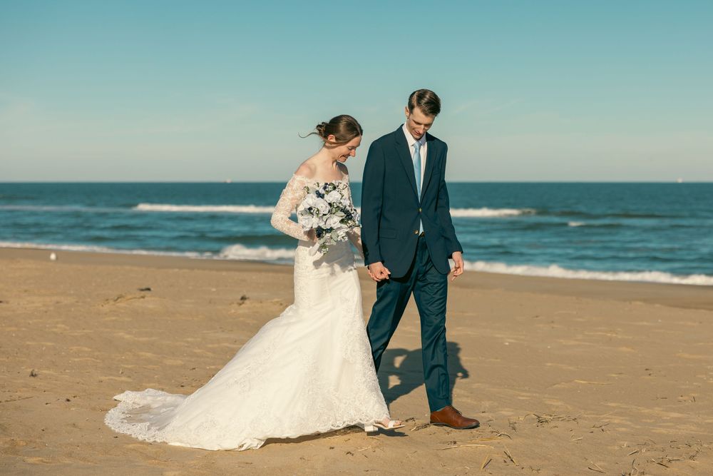 Bride and groom walking hand in hand on a sandy beach with ocean waves in the background on their wedding day.