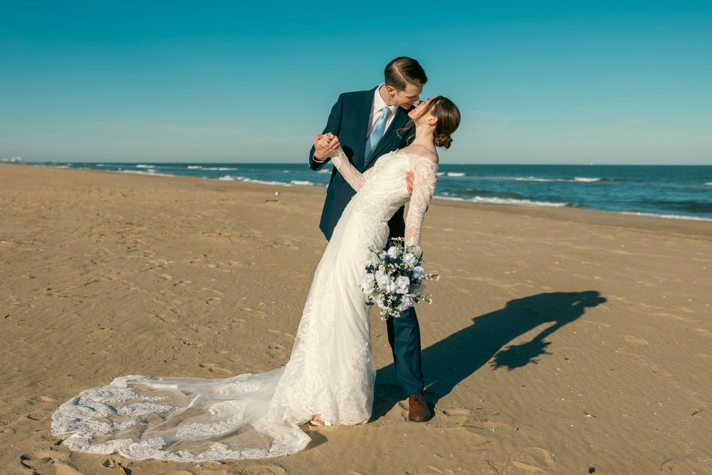 Bride and groom sharing a passionate kiss on a sunny beach, bride holding a blue and white floral bouquet.