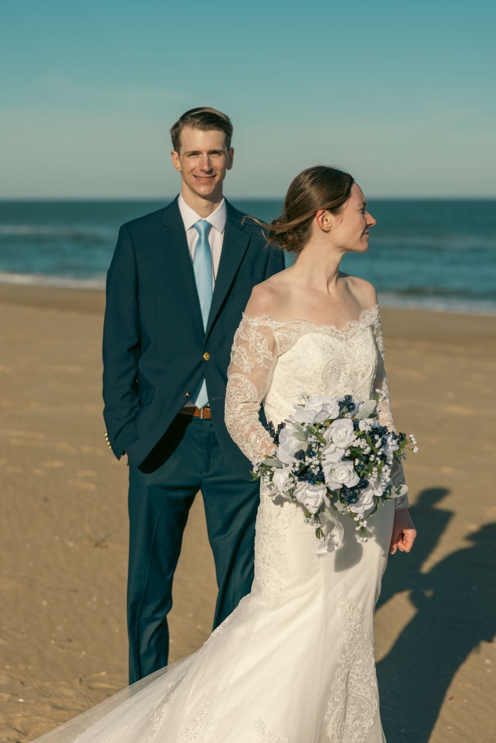 Elegant beach wedding portrait of bride in lace gown holding bouquet while groom in navy suit smiles at camera.