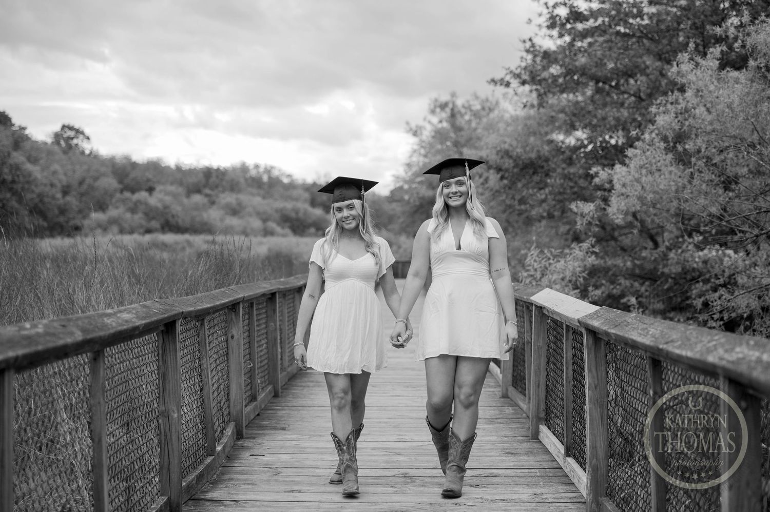 Two graduates in matching white dresses and graduation caps walk together on a wooden boardwalk path.