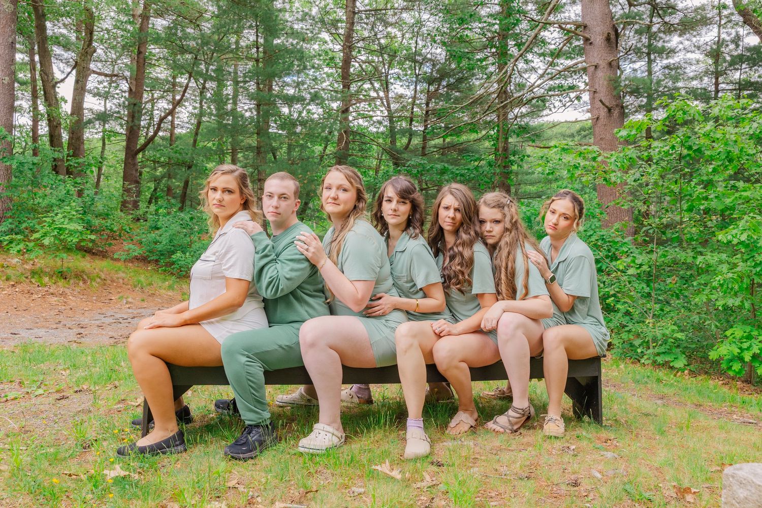 A group wearing matching sage green outfits sits together on a wooden bench in a forest setting.