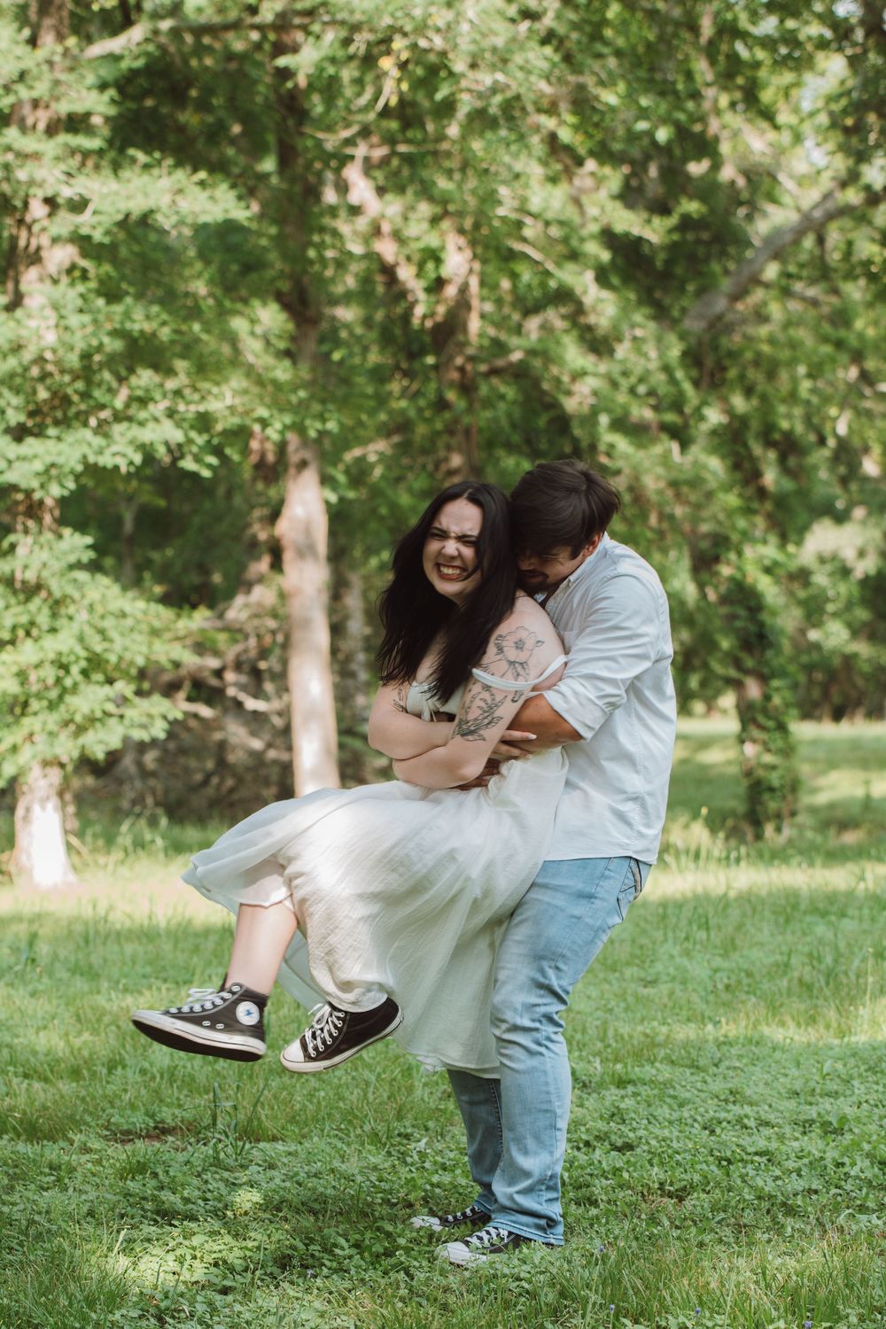 A couple in casual clothing shares a playful embrace while standing on lush green grass in a sunlit park setting.