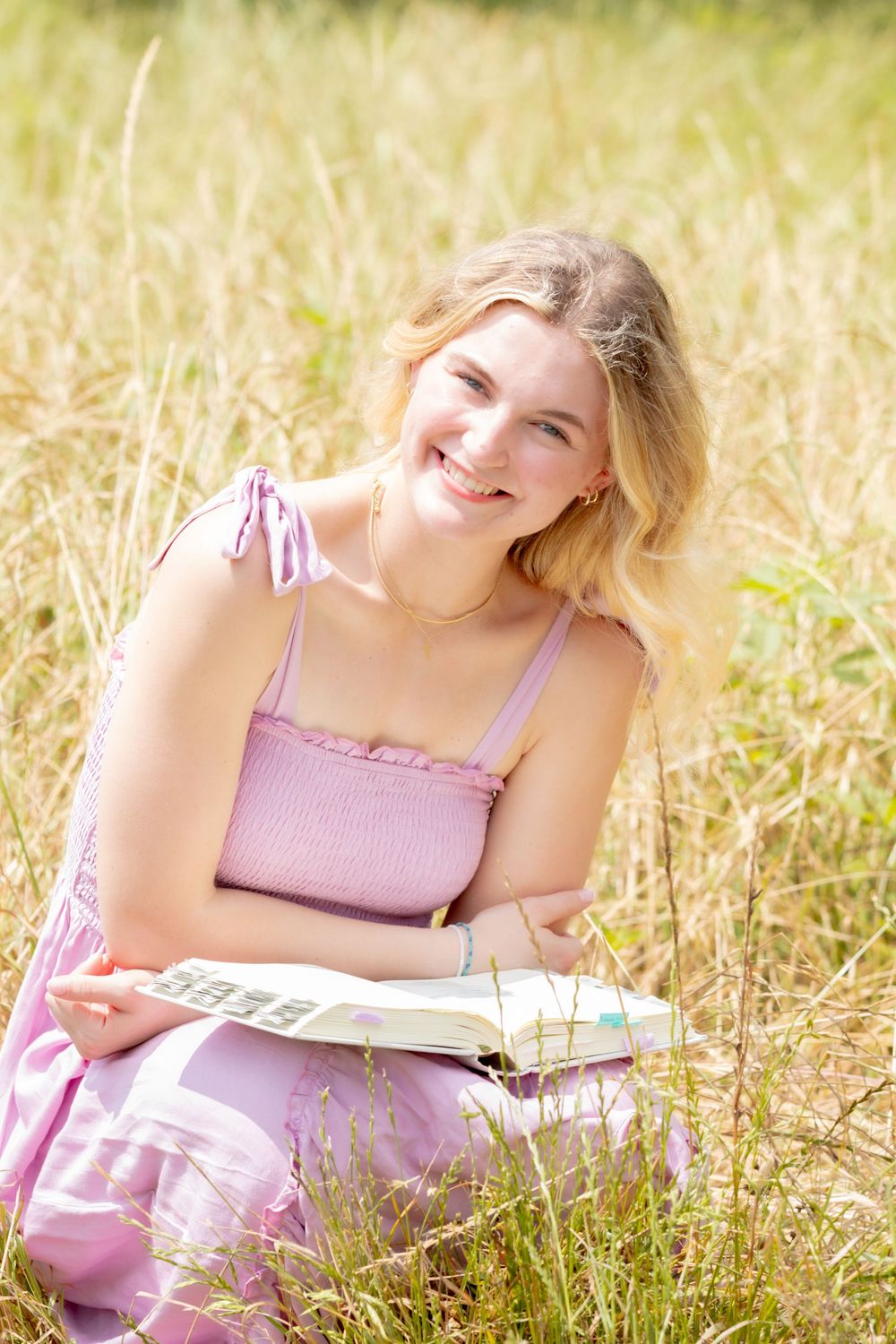 A figure in a pink dress reads a book while lying in a sun-drenched meadow of golden dried grass.
