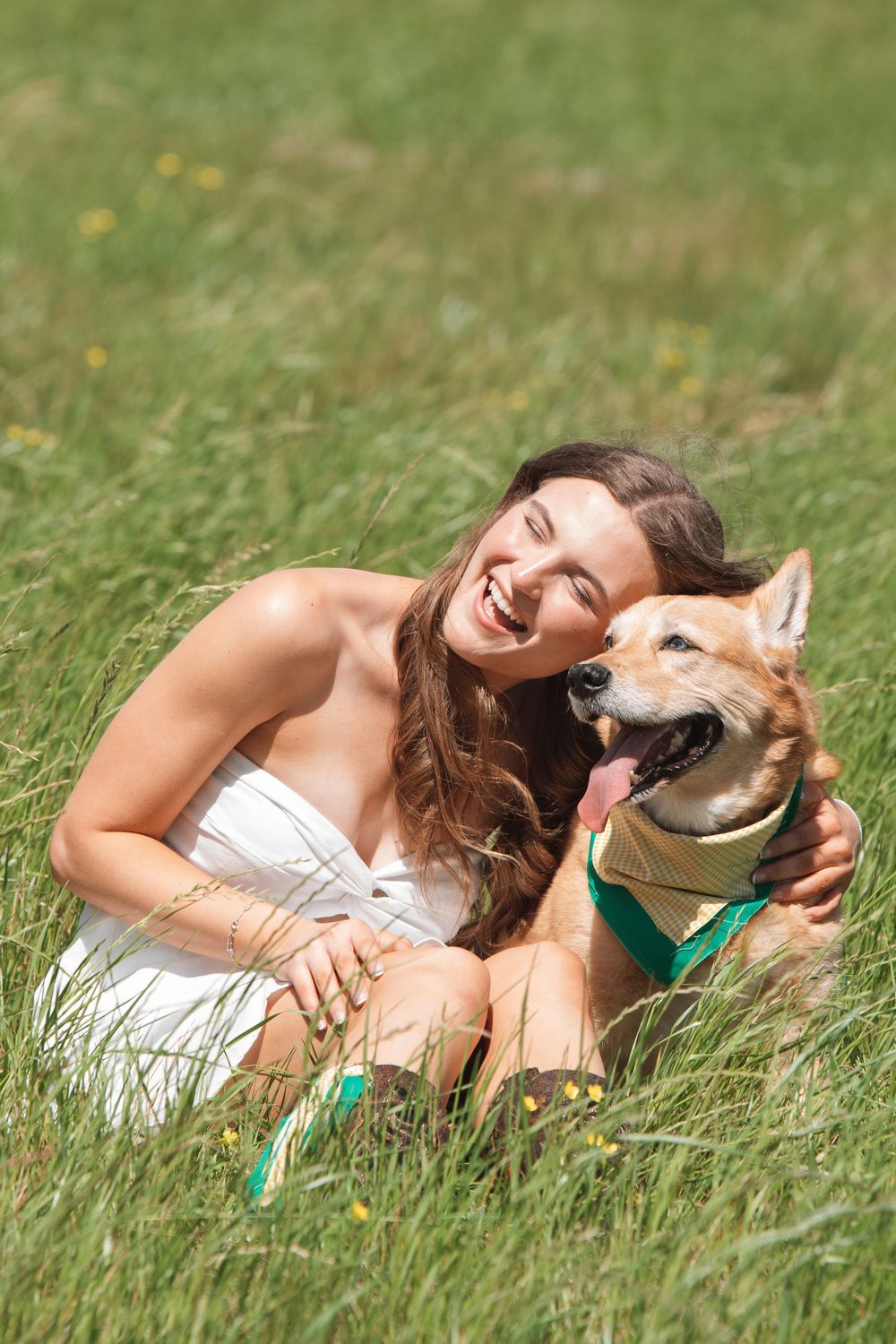 Person in white dress sharing joyful moment with happy dog wearing green bandana while sitting in grassy meadow.