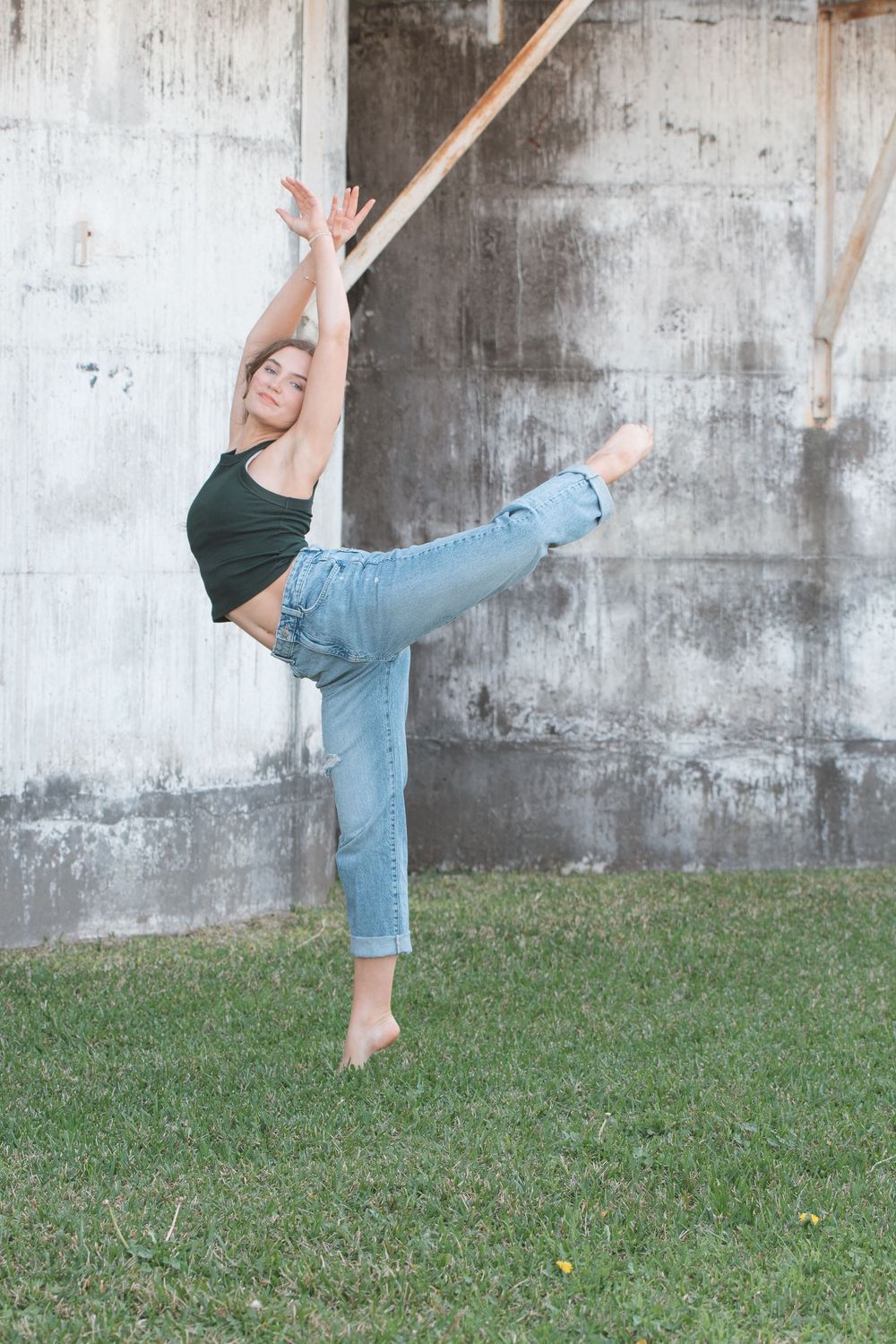 Dancer in casual wear performs high kick against concrete wall on grassy area outdoors.