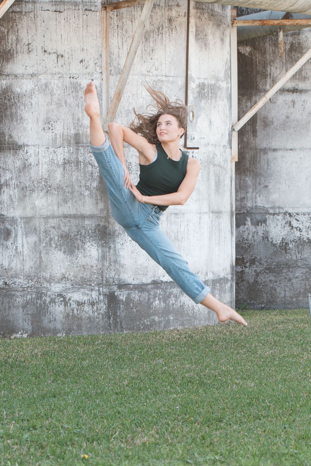 A dancer in a black top and blue pants performs a dynamic leap against a concrete wall and grass background.
