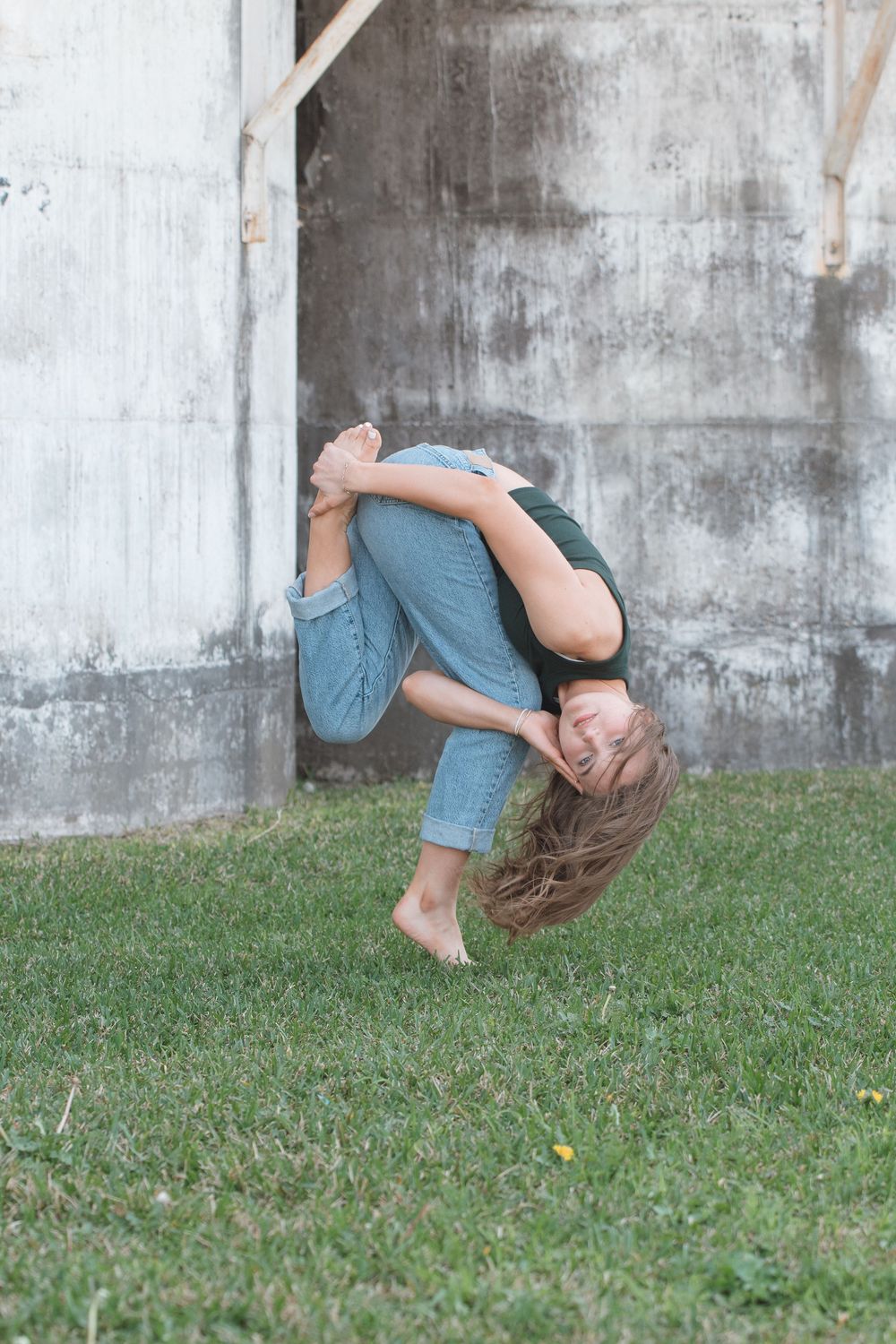 A person in denim outfit curls into a playful ball pose while standing barefoot on grass against a concrete wall.