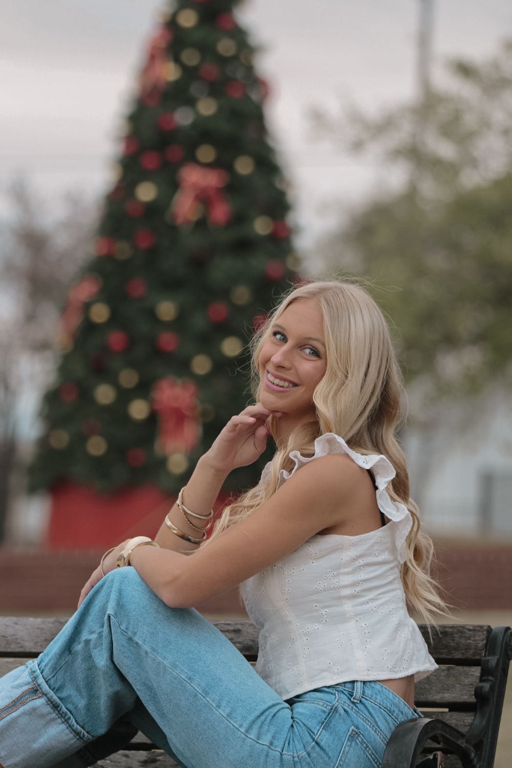 Person in white top and jeans poses on bench with decorated Christmas tree in background.