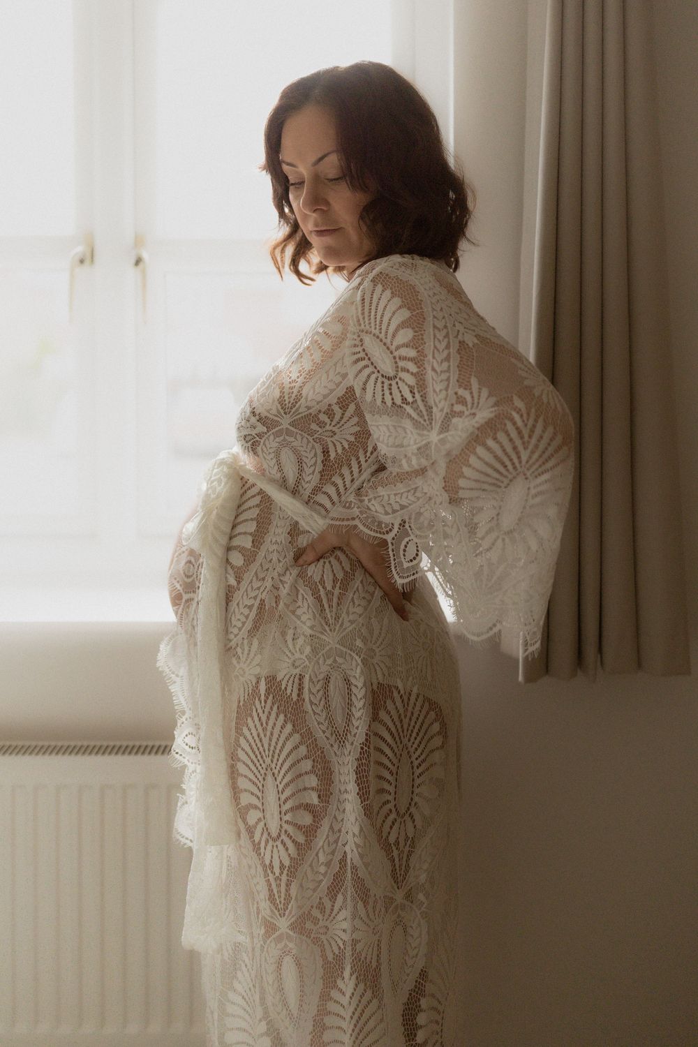 A person poses in a delicate white lace robe with intricate leafy patterns near a sunlit window.
