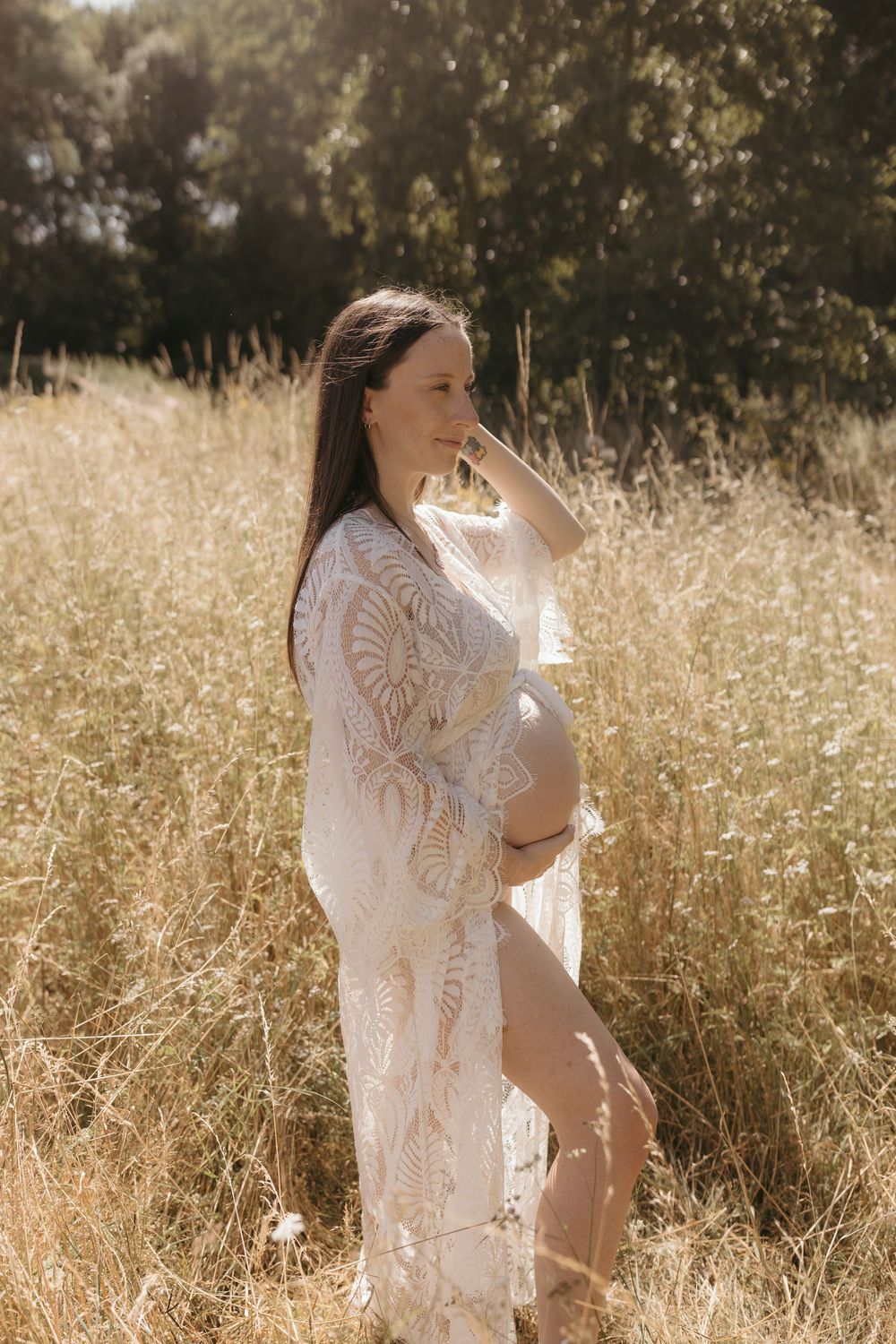 An expectant mother in a white lace dress stands gracefully in a sunlit field of golden grass.