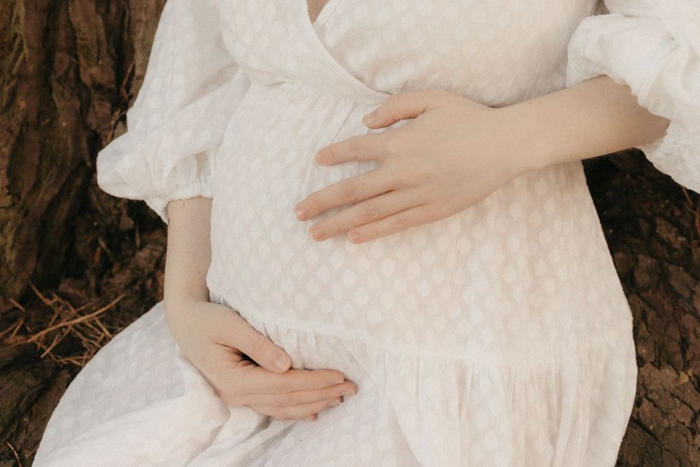 A person wearing a white dress holds a baby swaddled in white fabric while standing outdoors in nature.