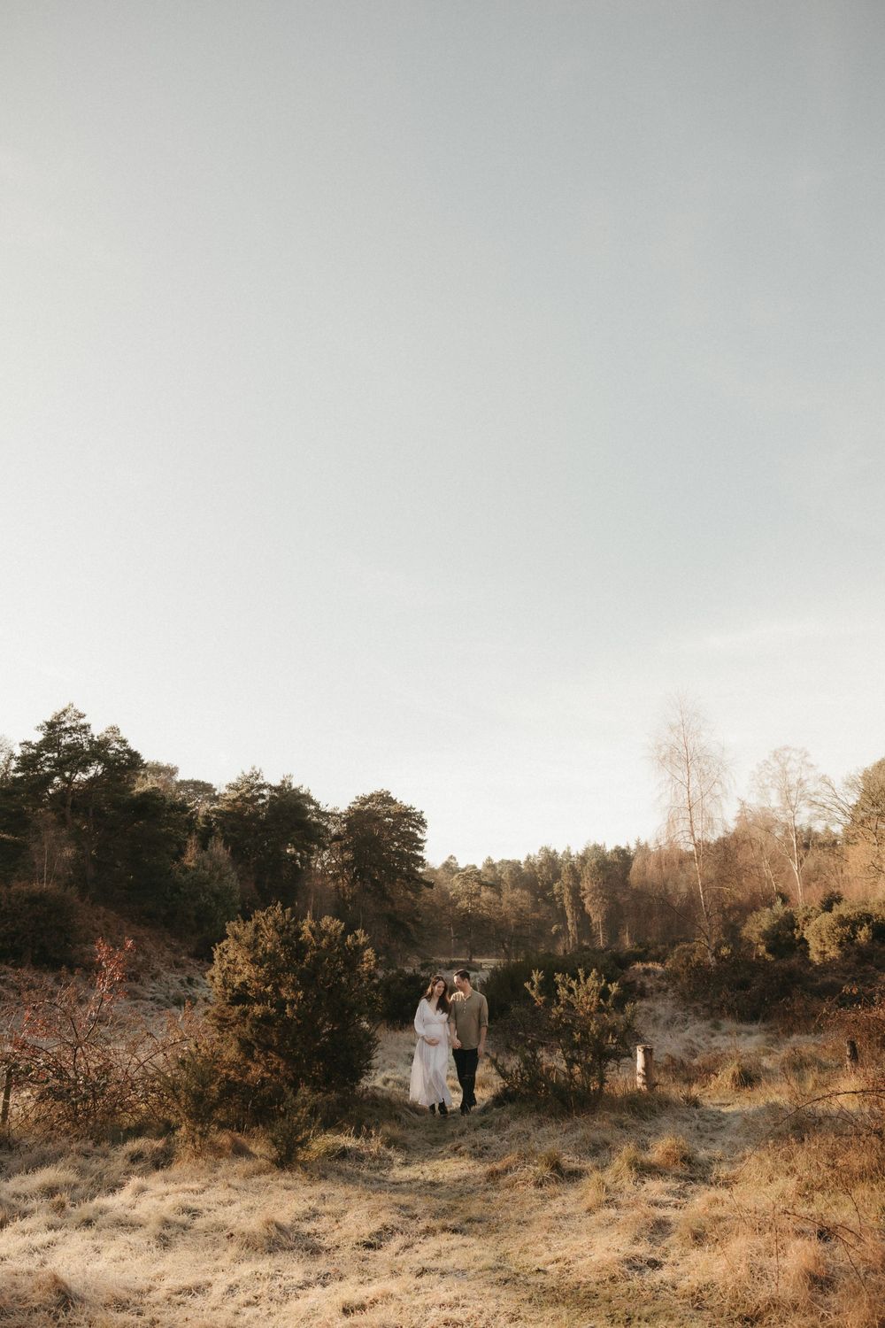 A couple stands in a golden field at sunset surrounded by desert brush and juniper trees in a romantic outdoor setting.