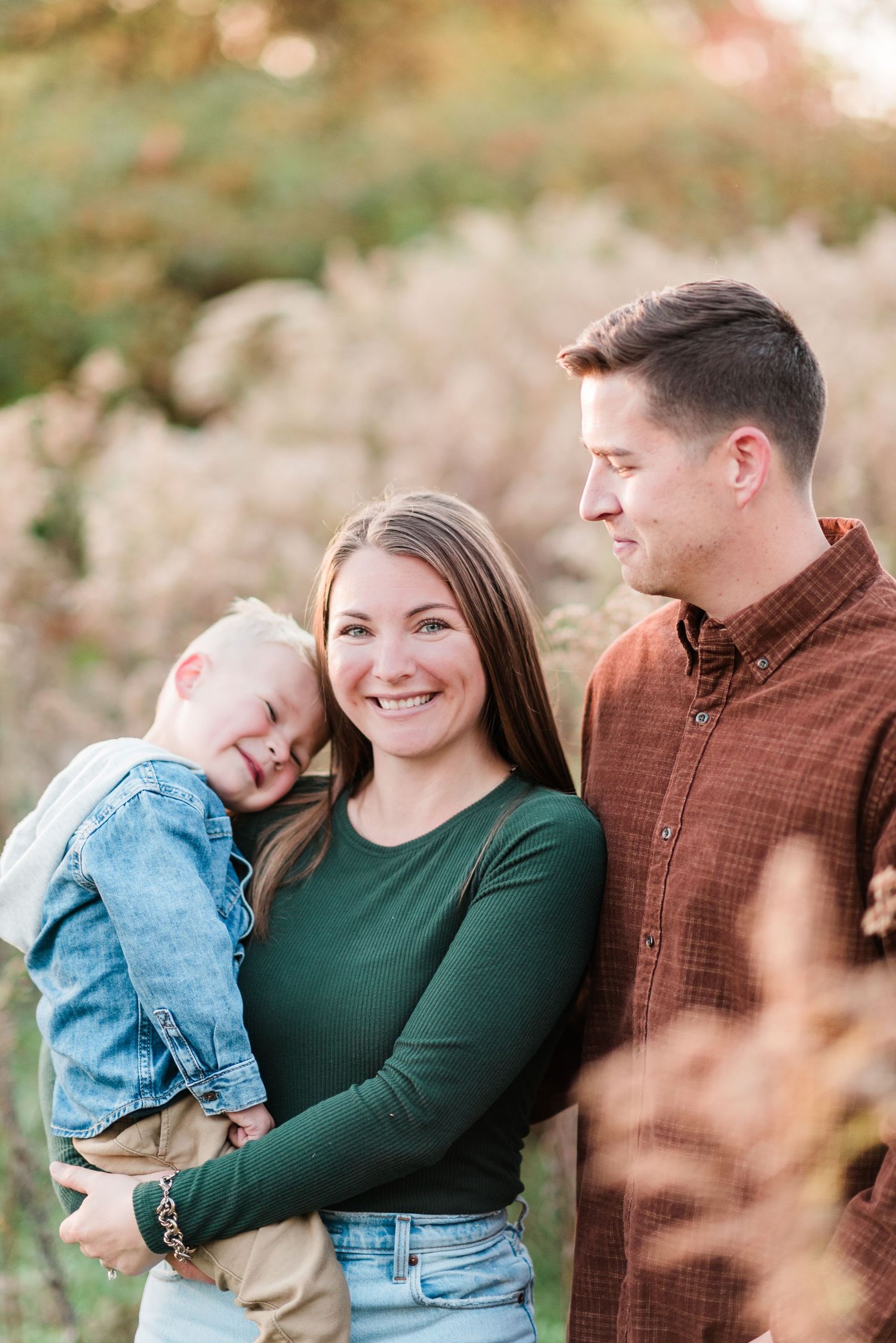 A family of three poses for an outdoor autumn portrait surrounded by soft golden bokeh and foliage.