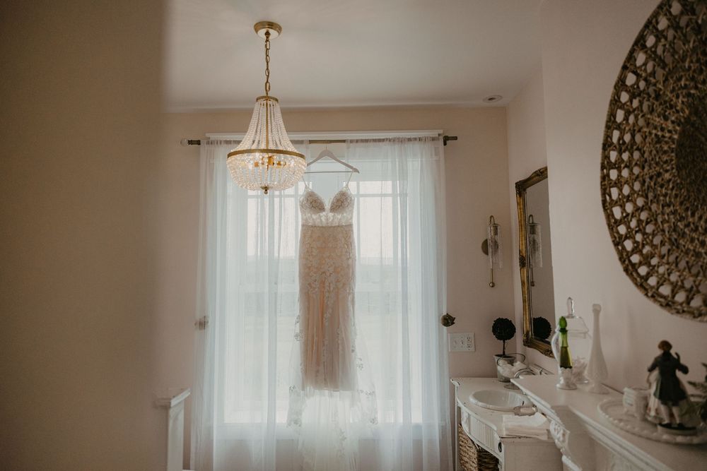 A wedding dress hangs elegantly from a chandelier in a bright room with sheer curtains and decorative wall art.