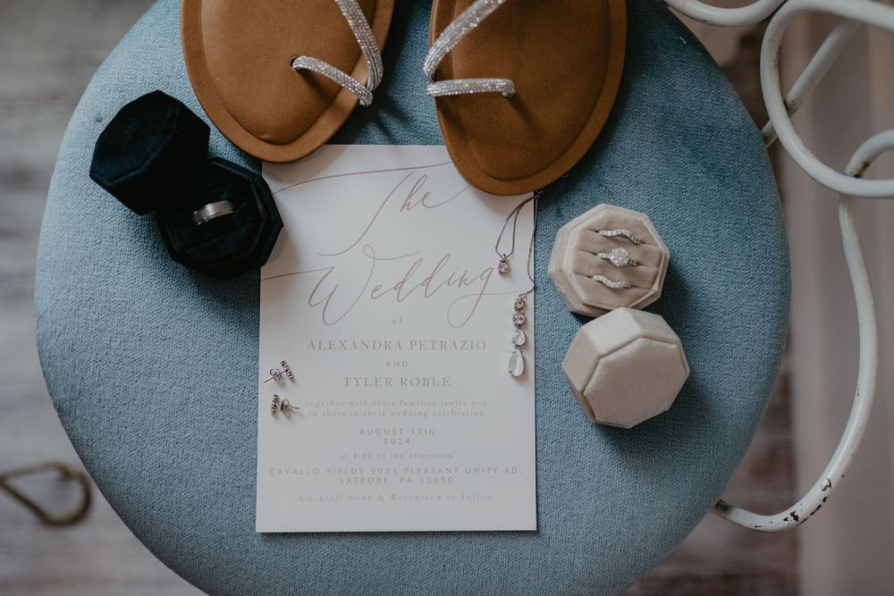Wedding invitation card displayed with brown leather shoes, black box and decorative items on a light blue surface.