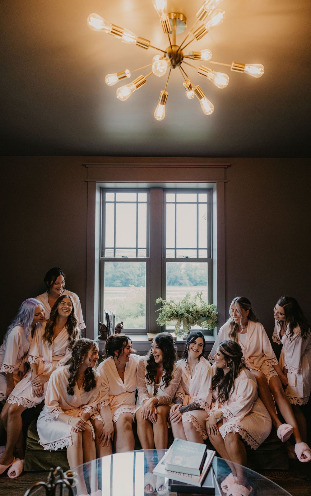 Bridal party in matching robes gathers by window beneath sputnik chandelier during wedding preparations.