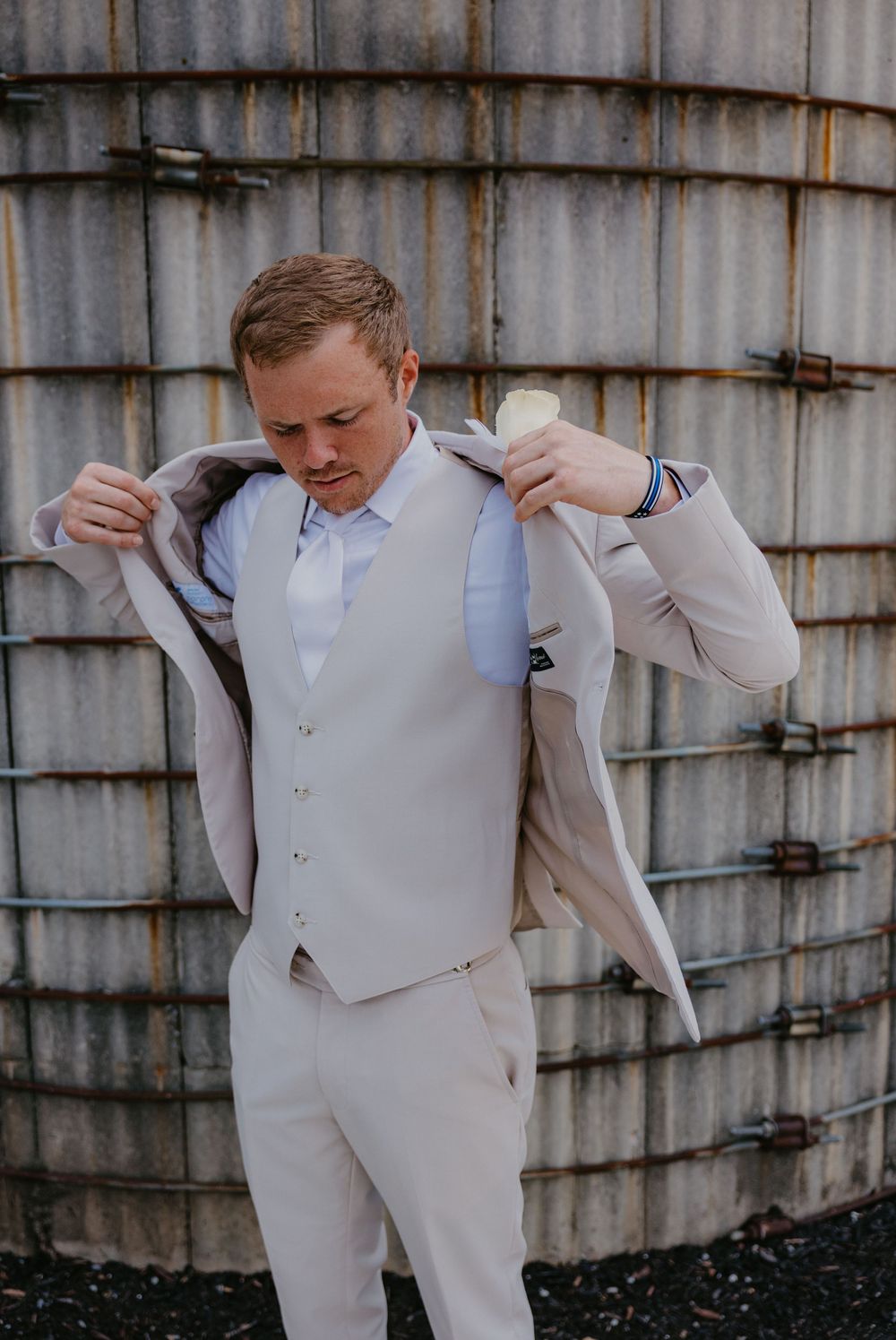 Someone in a light gray suit and vest putting on a formal jacket against an industrial metal wall backdrop.