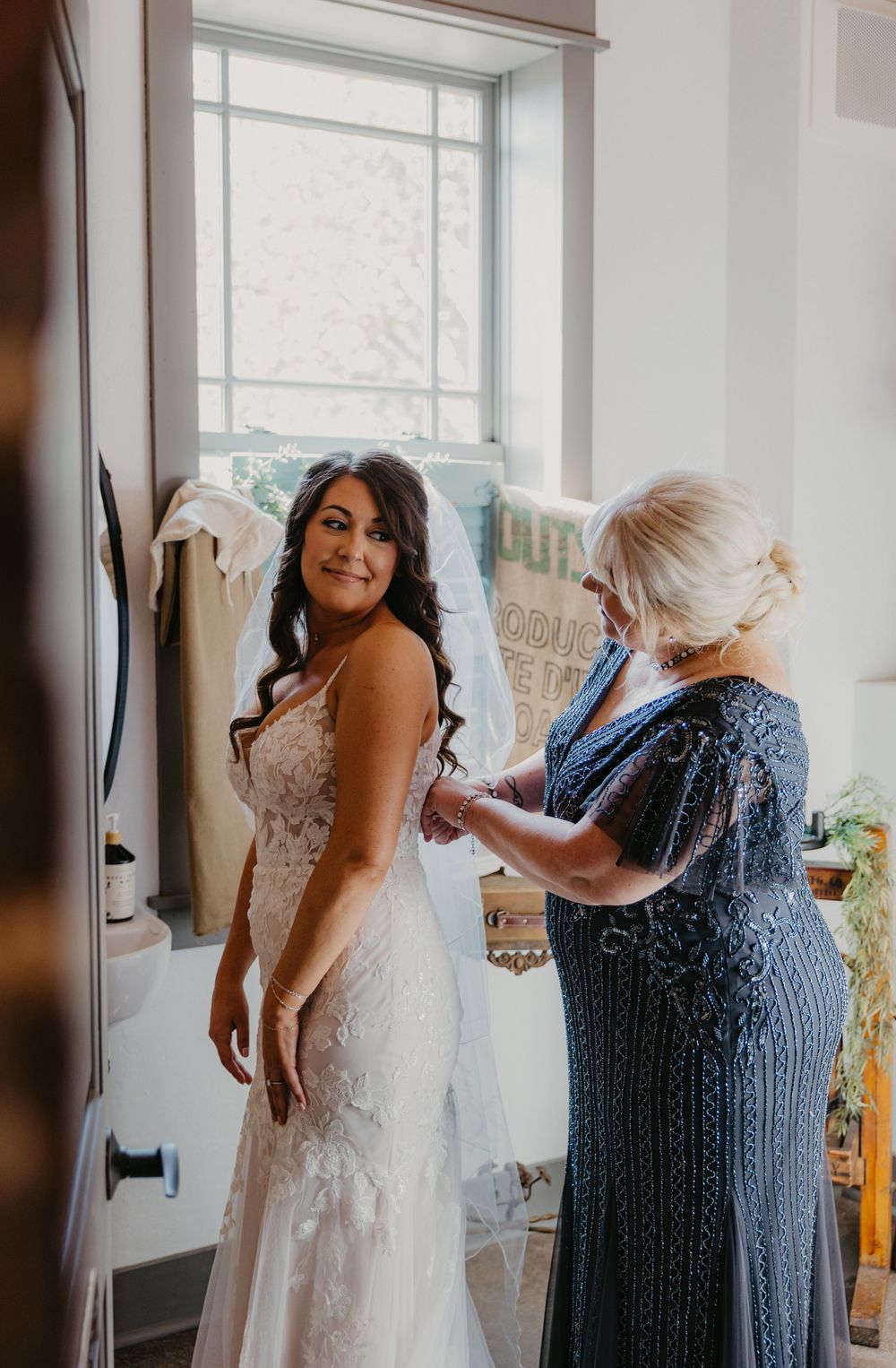 An elegant lace wedding dress is being fitted in a sunlit room with soft window light streaming in.