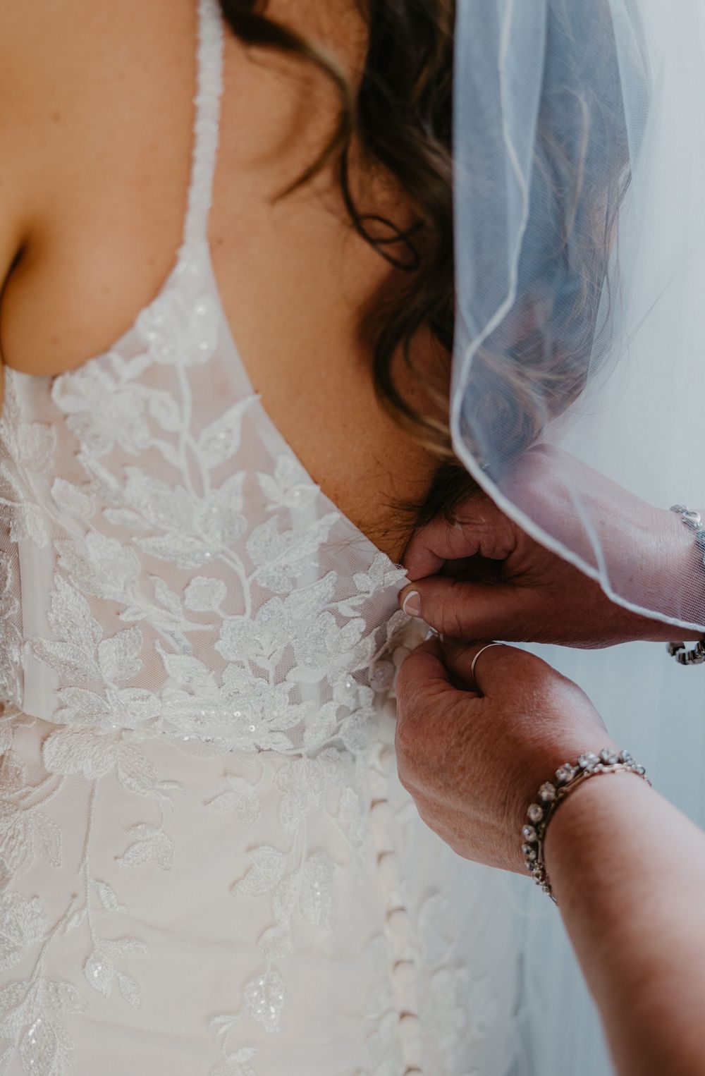 Close-up of hands fastening delicate lace buttons on a white wedding dress, with bridal veil visible in background.