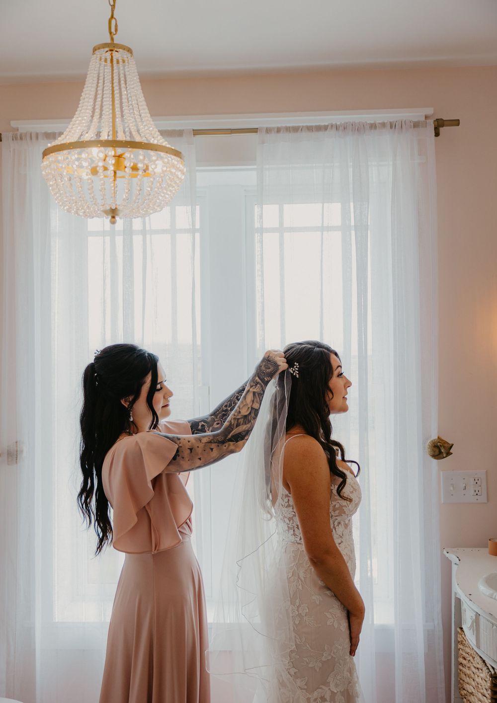 A bridesmaid in a blush pink dress helps adjust a wedding veil in a sunlit room with sheer curtains and crystal chandelier.