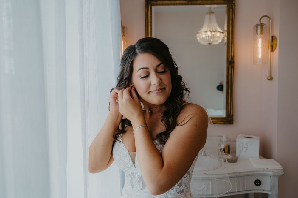 A person in a white outfit putting on jewelry while standing by a vanity with a mirror and sconce lighting.