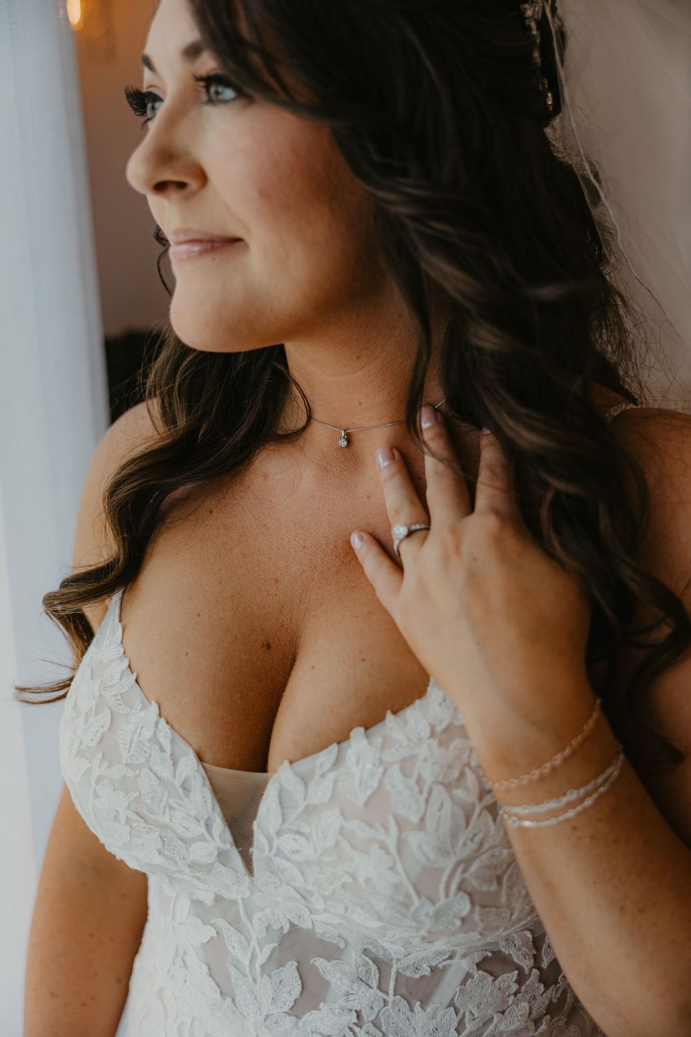 A close-up view of a bride in a white lace wedding dress with long dark curled hair touching her necklace.