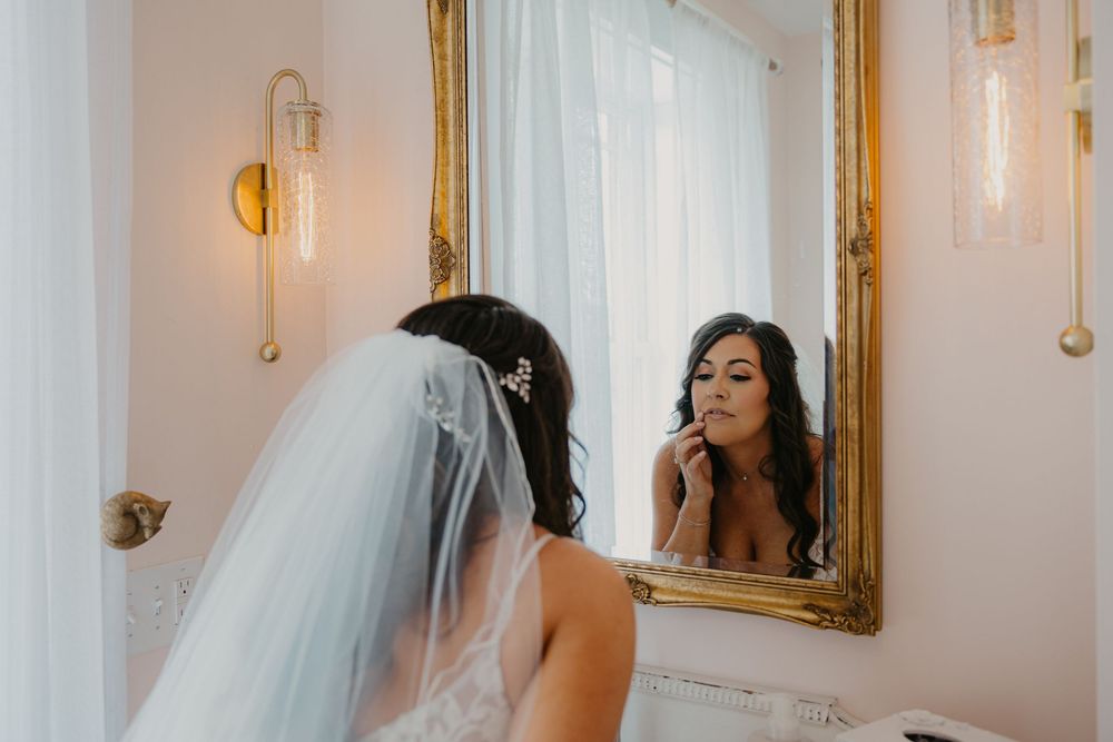 Reflection in an ornate gold mirror shows an elegant figure in flowing white veil preparing for a special occasion.