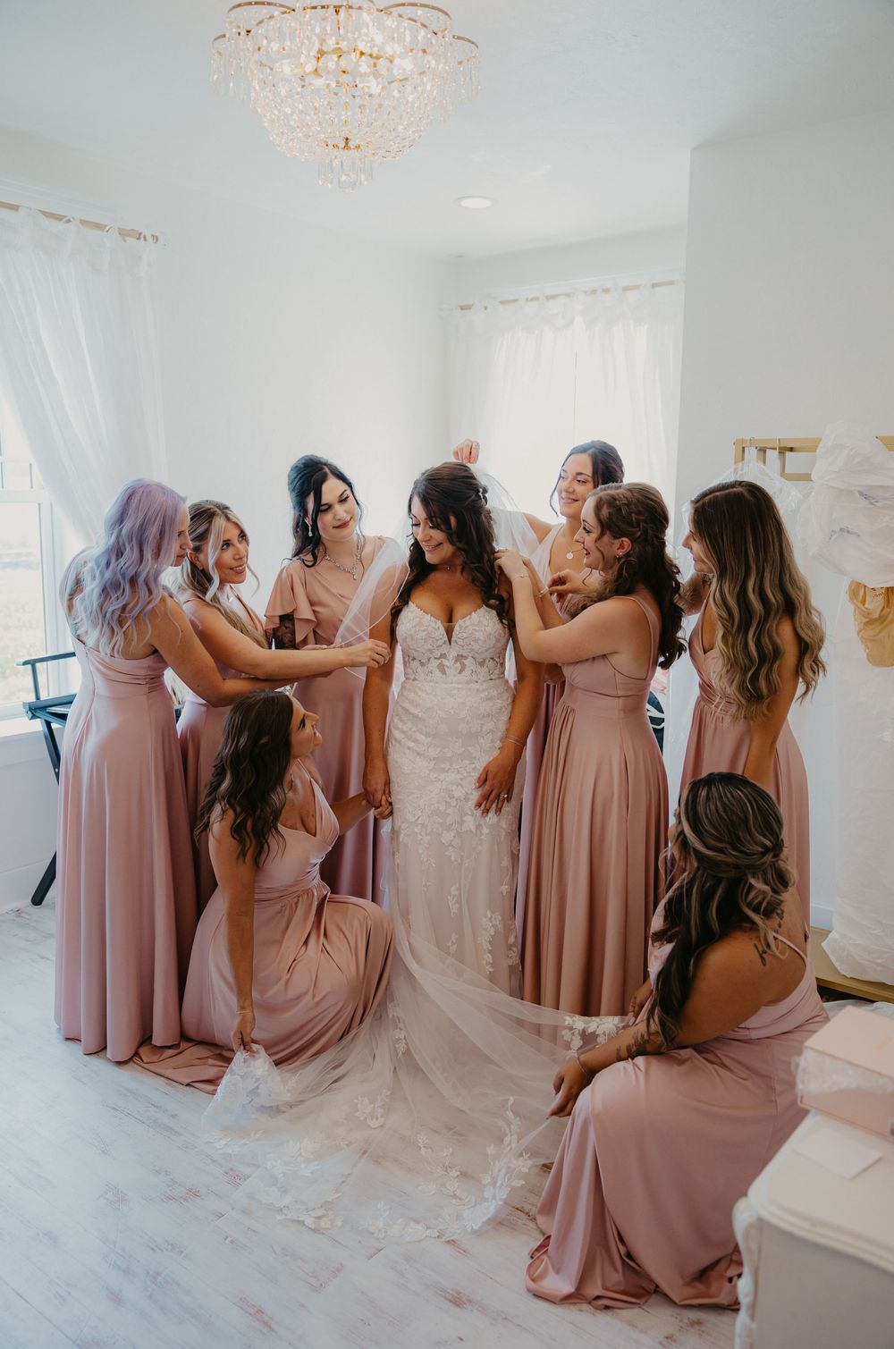 A group in blush pink bridesmaid dresses gathers around a bride in a white lace gown in an elegant bright room.