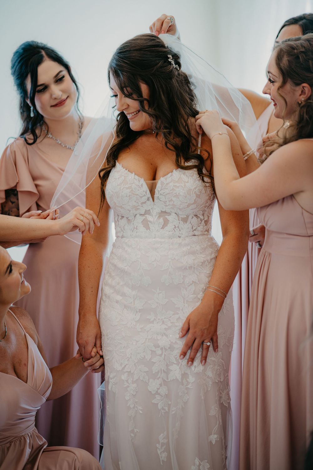 Bridesmaids in dusty pink dresses help adjust a bride's lace wedding gown during wedding preparations.