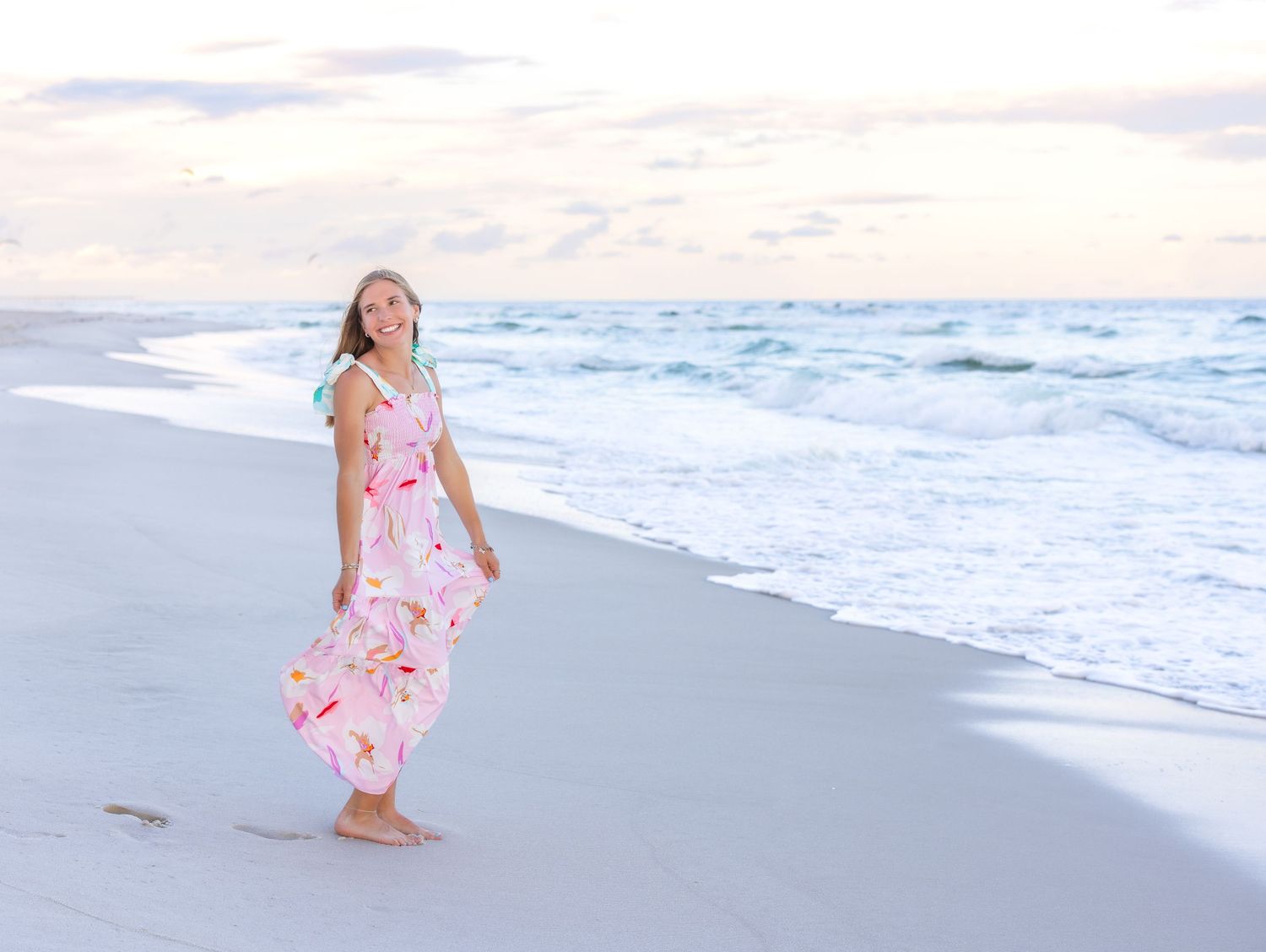 A person in a pink floral dress walks along the beach at sunset while ocean waves roll onto shore.