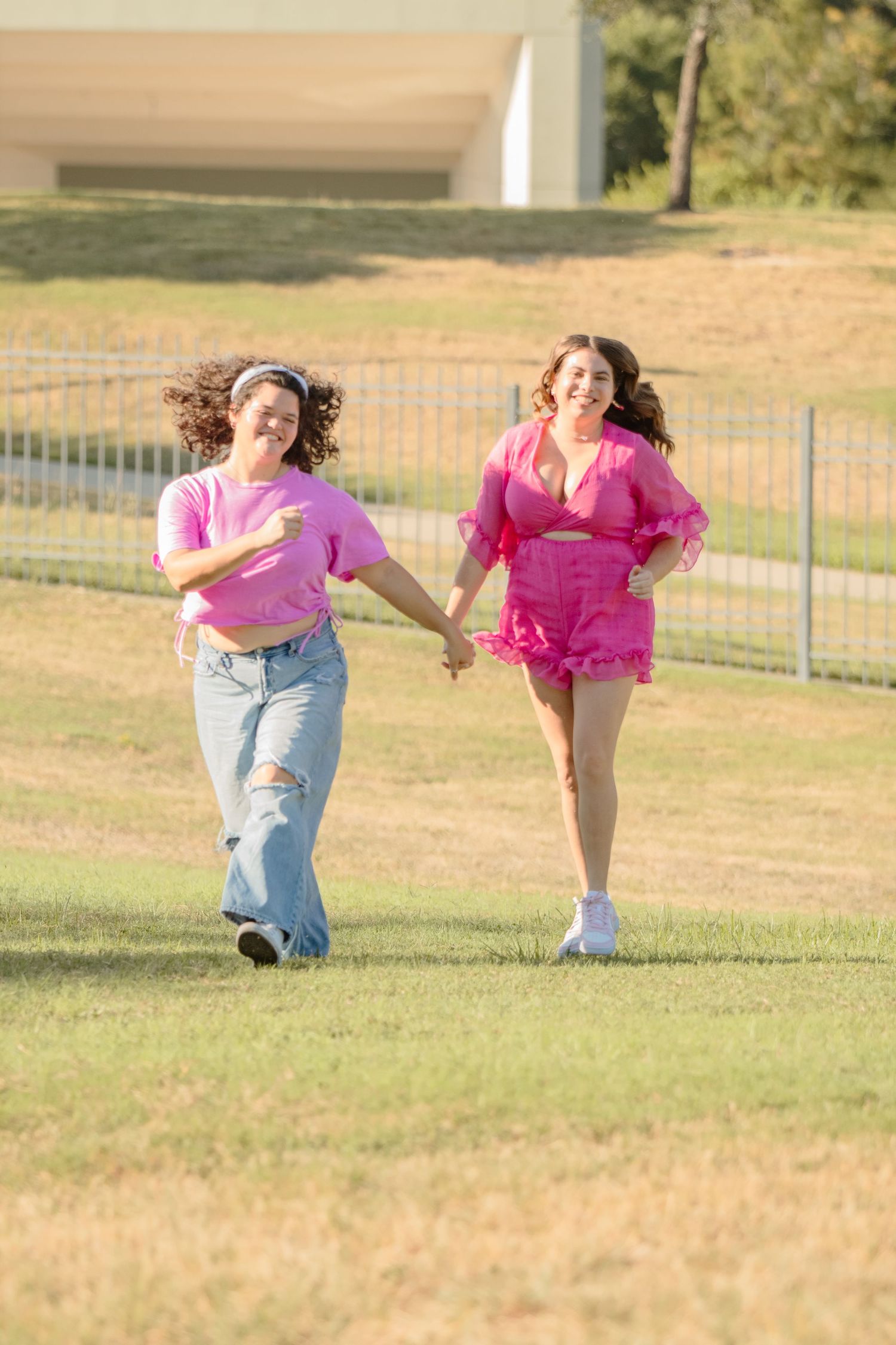Two young dancers in pink costumes practicing joyful leaps and dance moves on a grassy outdoor field.
