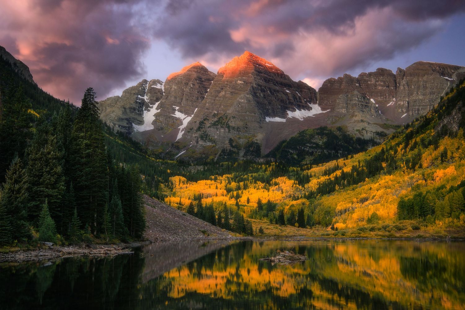 Maroon Bells mountains reflect in a lake during autumn with golden aspen trees and dramatic sunset clouds in Colorado.