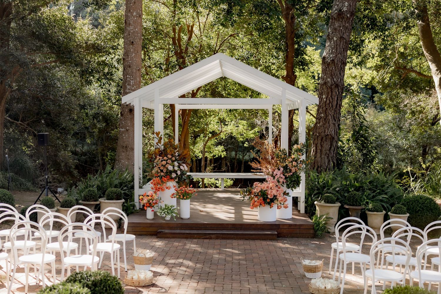 A white wedding gazebo surrounded by trees with rows of clear modern chairs on a brick patio for an outdoor ceremony.