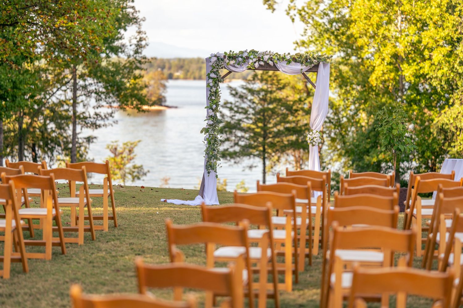 Wooden chairs arranged for an outdoor lakeside wedding ceremony with a floral arch and white fabric draping.
