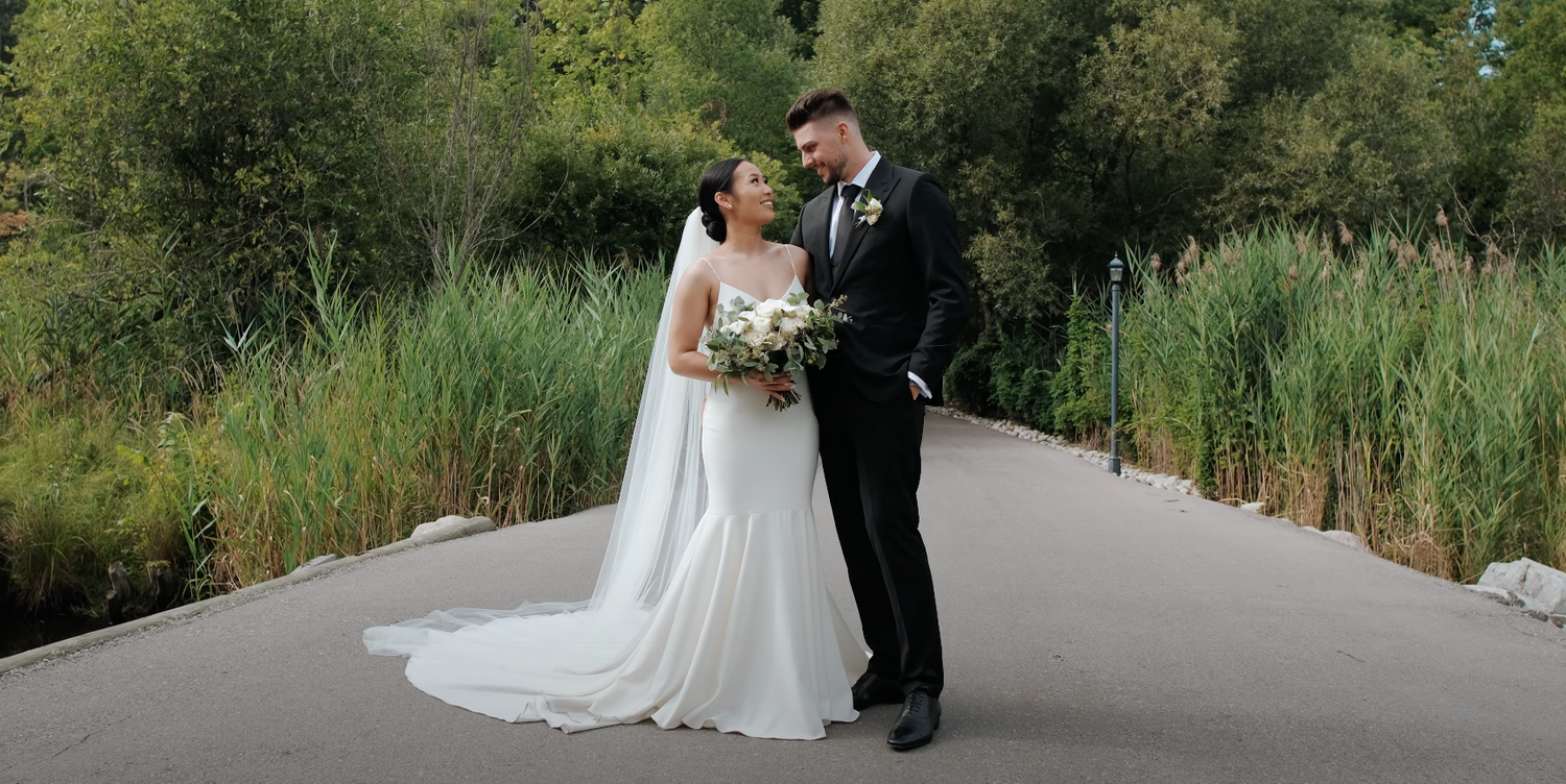 Bride in flowing white gown and groom pose on pathway surrounded by greenery.