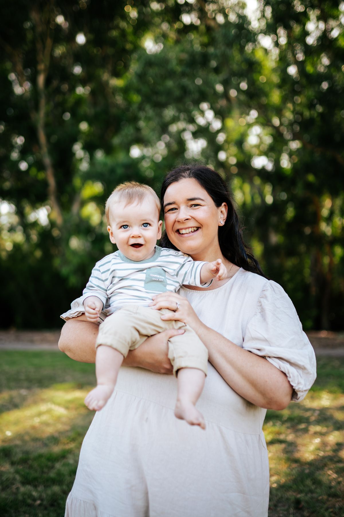 Woman holds and smiles with an adorable baby outdoors in a sunlit garden setting taken by Melbourne family photographer.