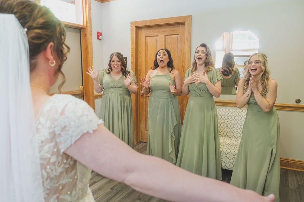 Bridesmaids in sage green dresses react emotionally in a getting-ready room.