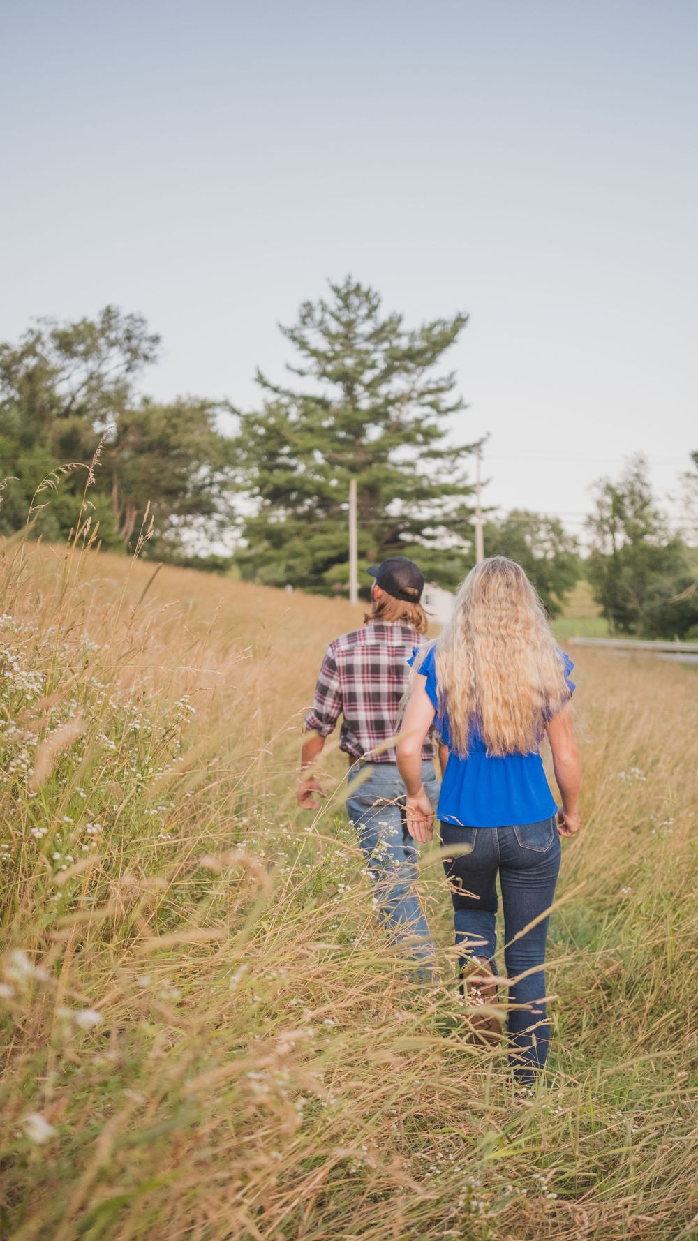A couple walking through tall grass on a hillside with pine trees in the distance.