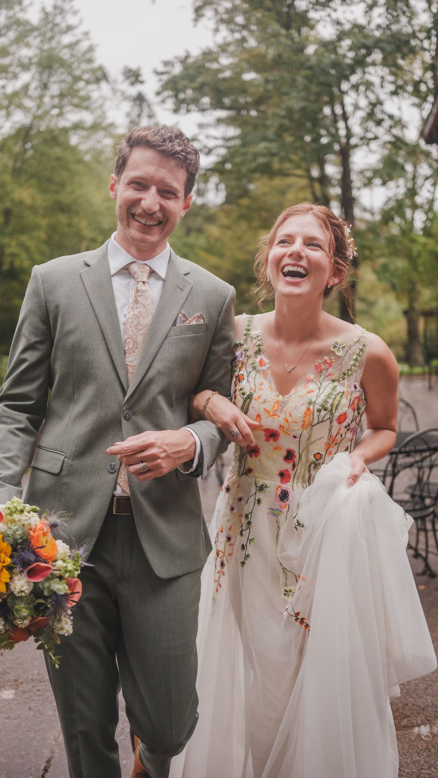 A joyful couple walking outdoors on their wedding day, wearing a grey suit and floral embroidered wedding gown.