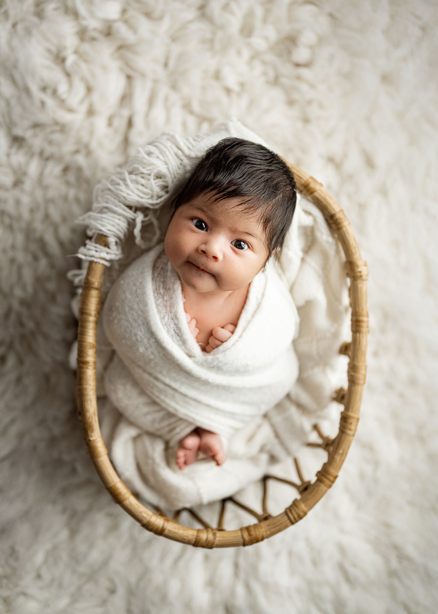 A newborn wrapped in white fabric lies in a circular woven basket against a cream background.