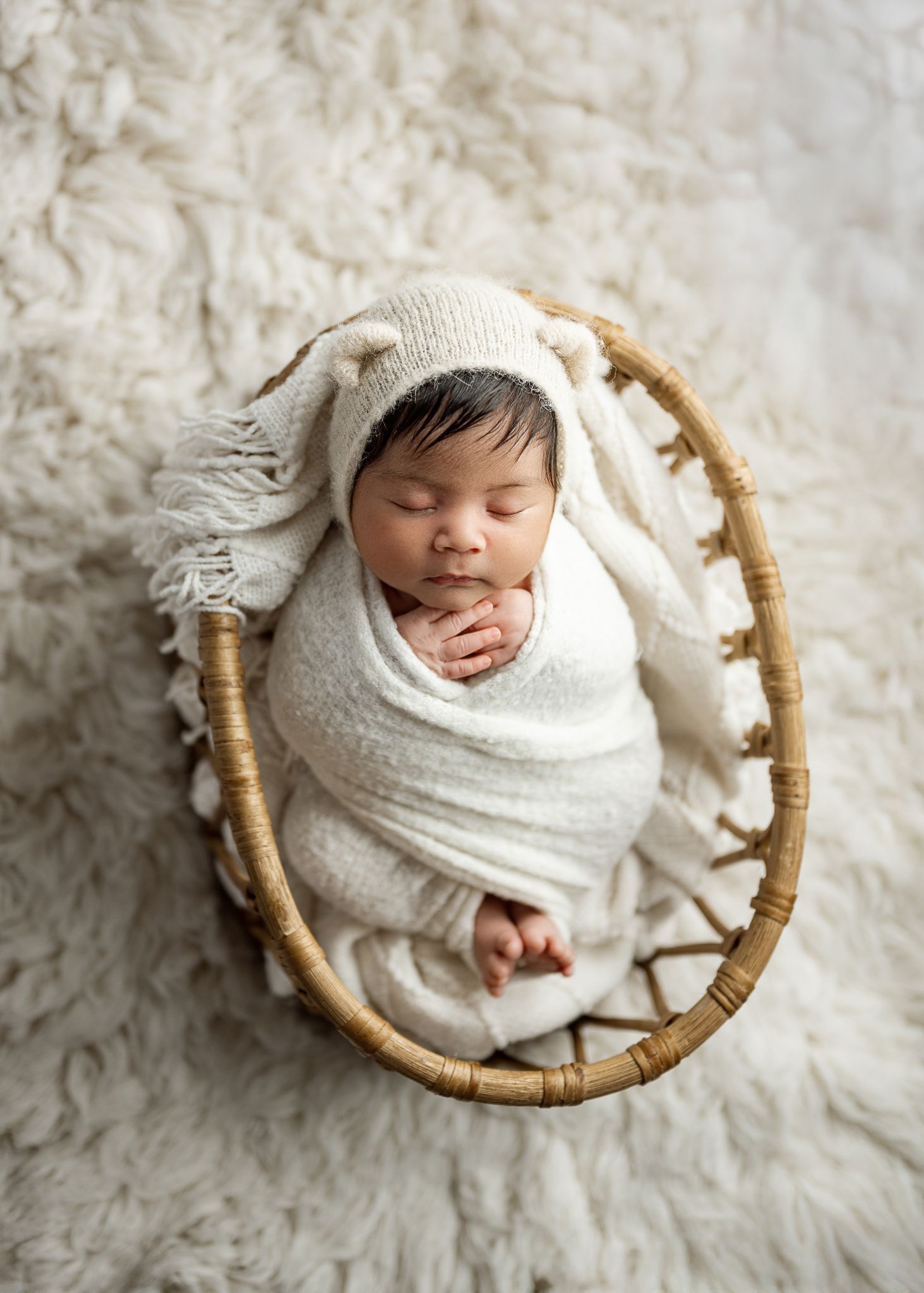A sleeping newborn baby wrapped in white swaddling cloth lies curled up in a round rattan basket on fluffy white fabric.