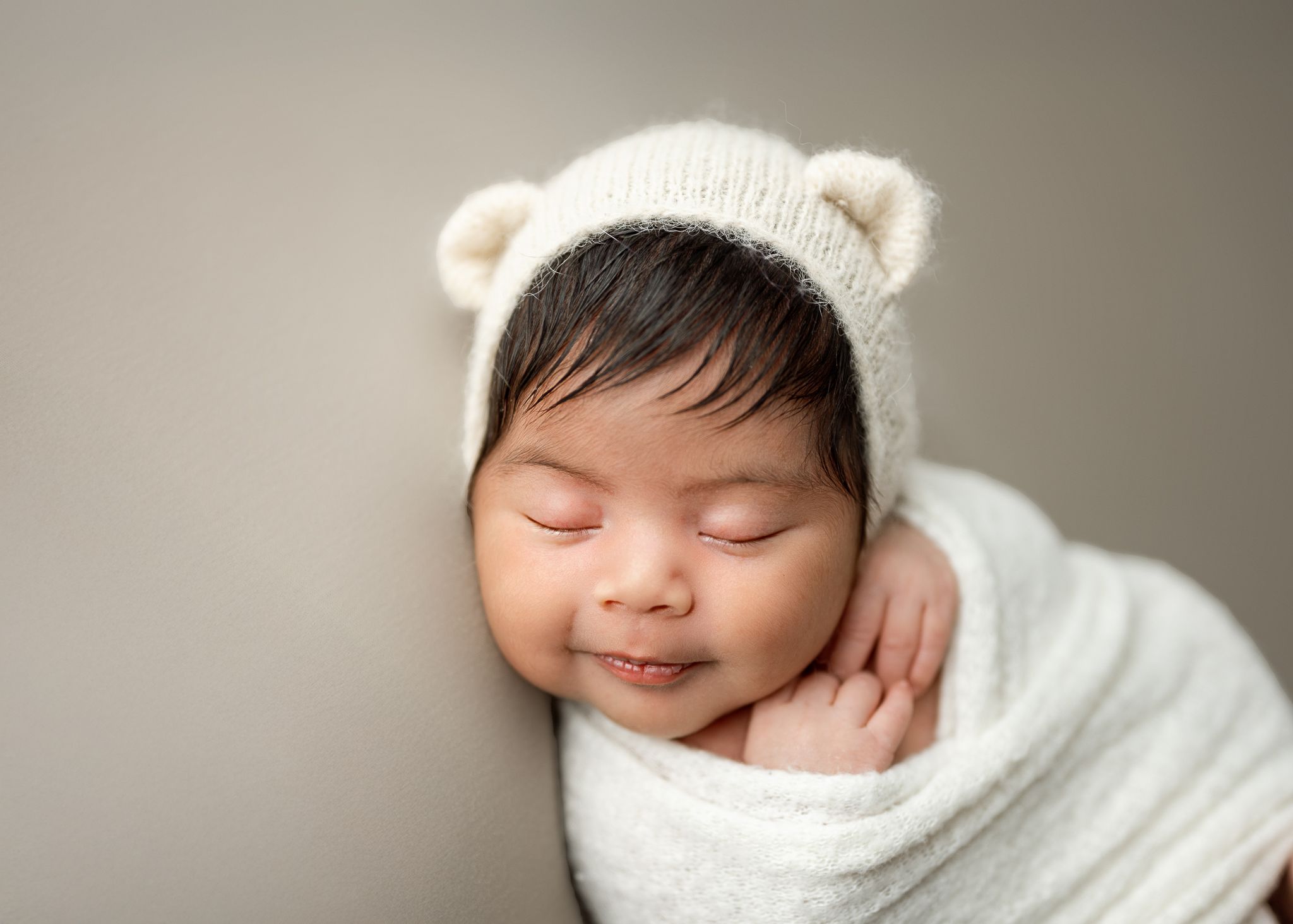 Sleeping newborn wearing white knit bear hat and wrapped in cozy white swaddle against neutral backdrop.