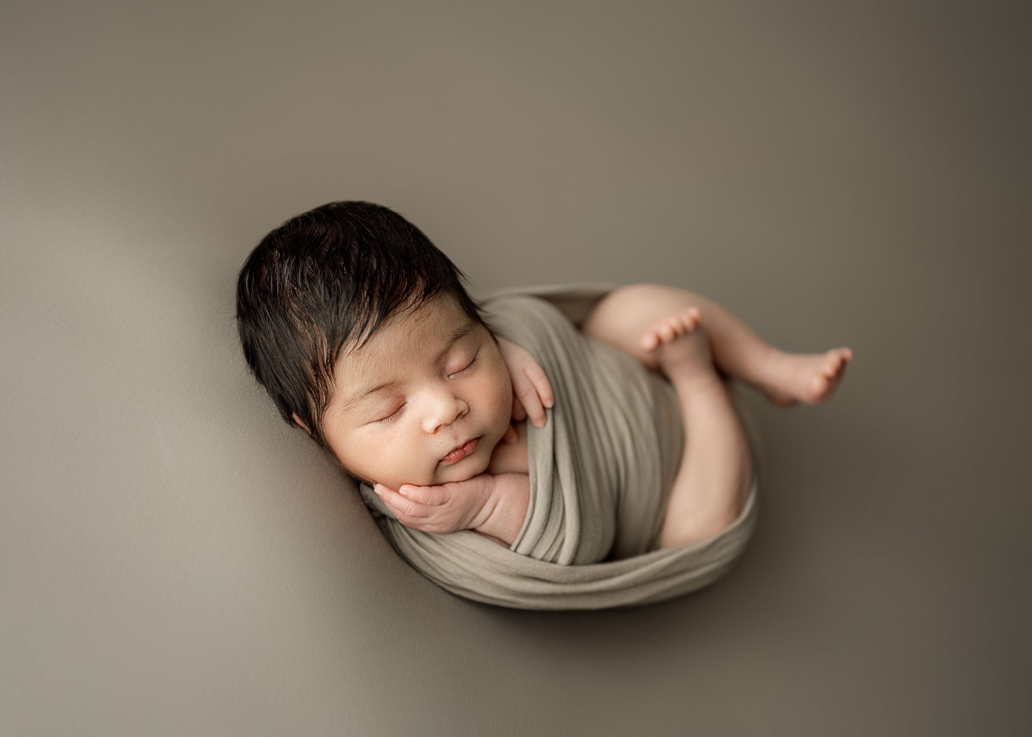 A peaceful sleeping newborn swaddled in soft beige fabric against a neutral backdrop.