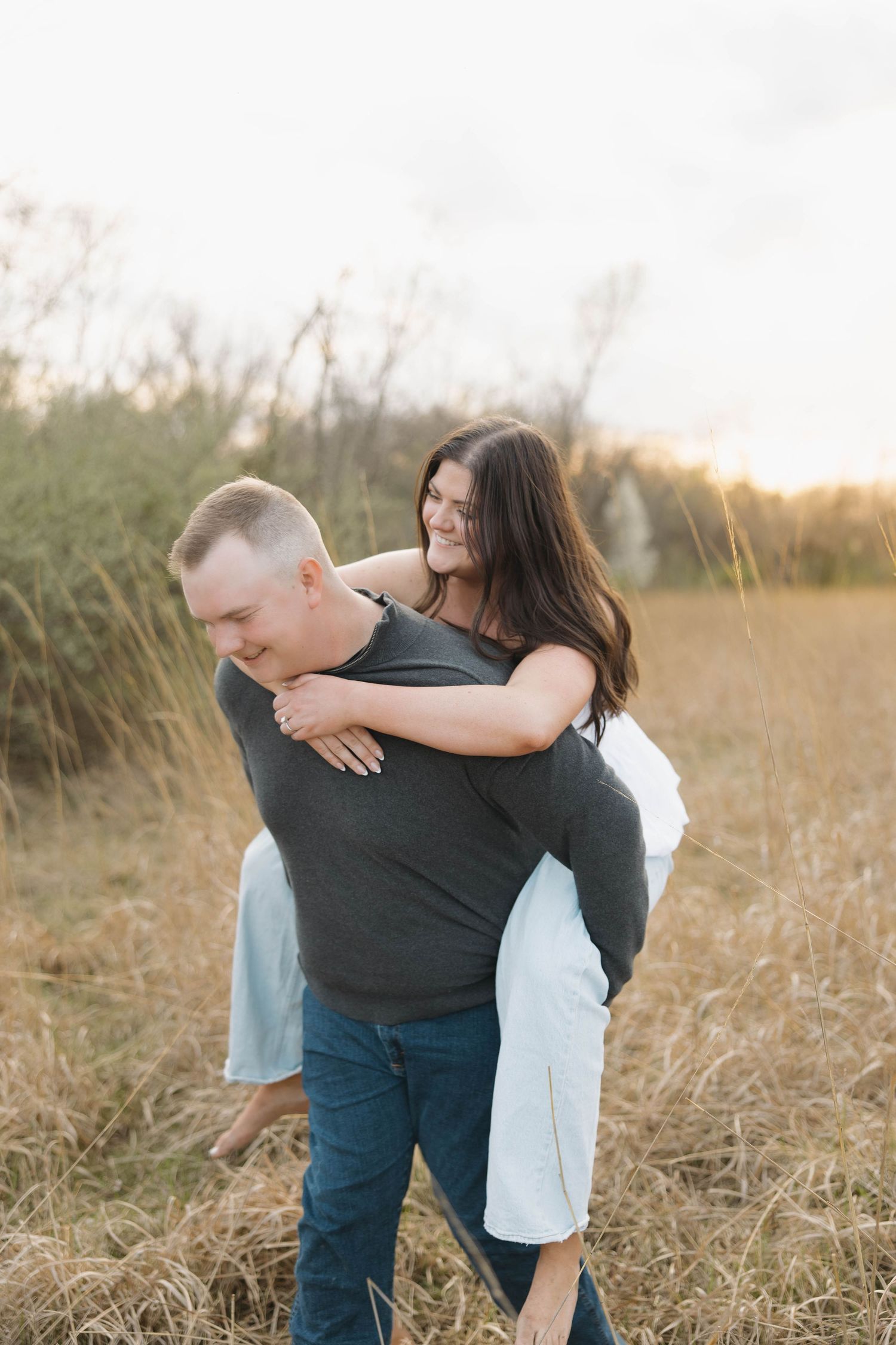 A couple embraces in a rustic field at sunset while wearing casual clothing during an outdoor photoshoot.