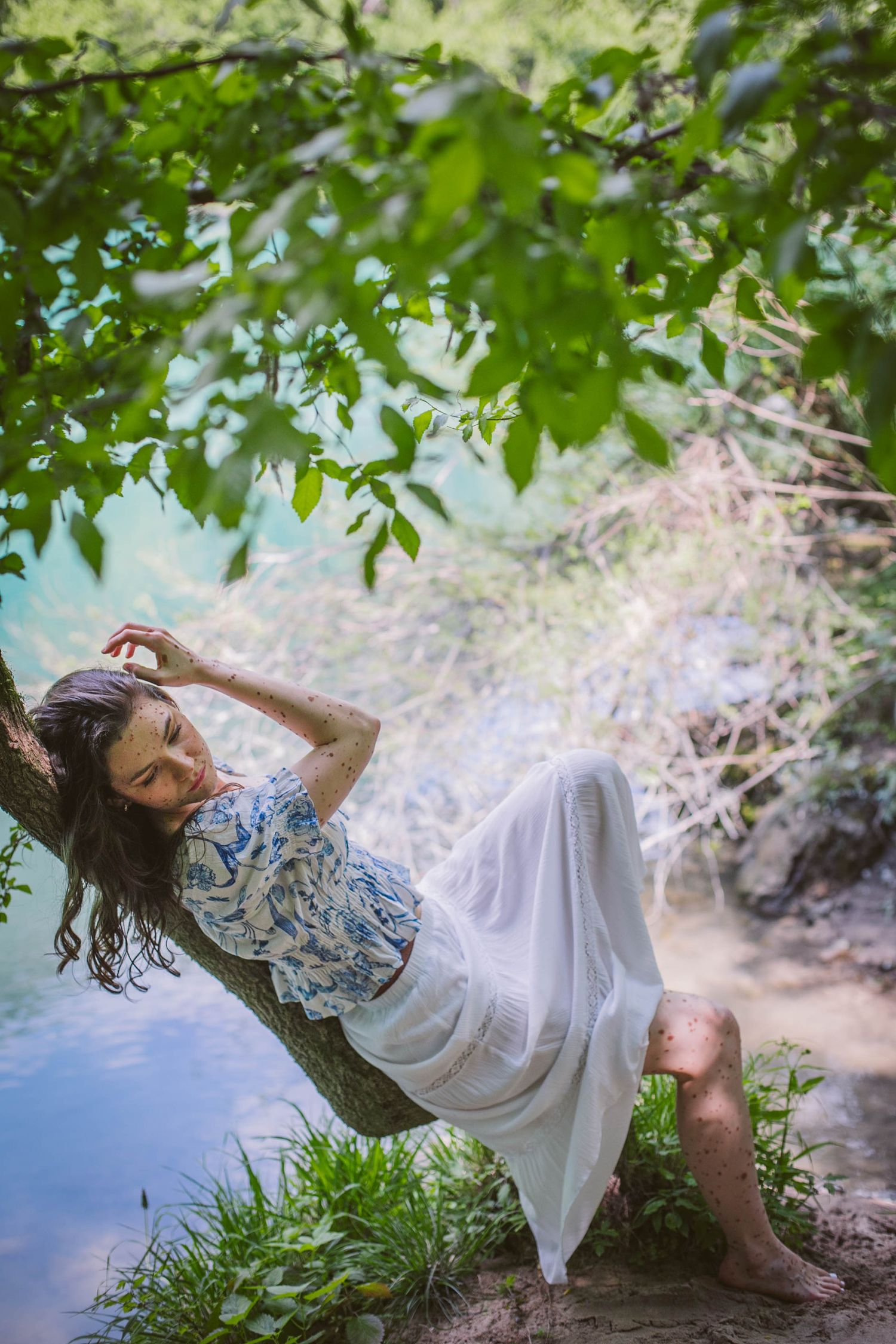 A dreamy portrait by a lake with flowing white dress and floral top as leaves create natural framing against blue water.