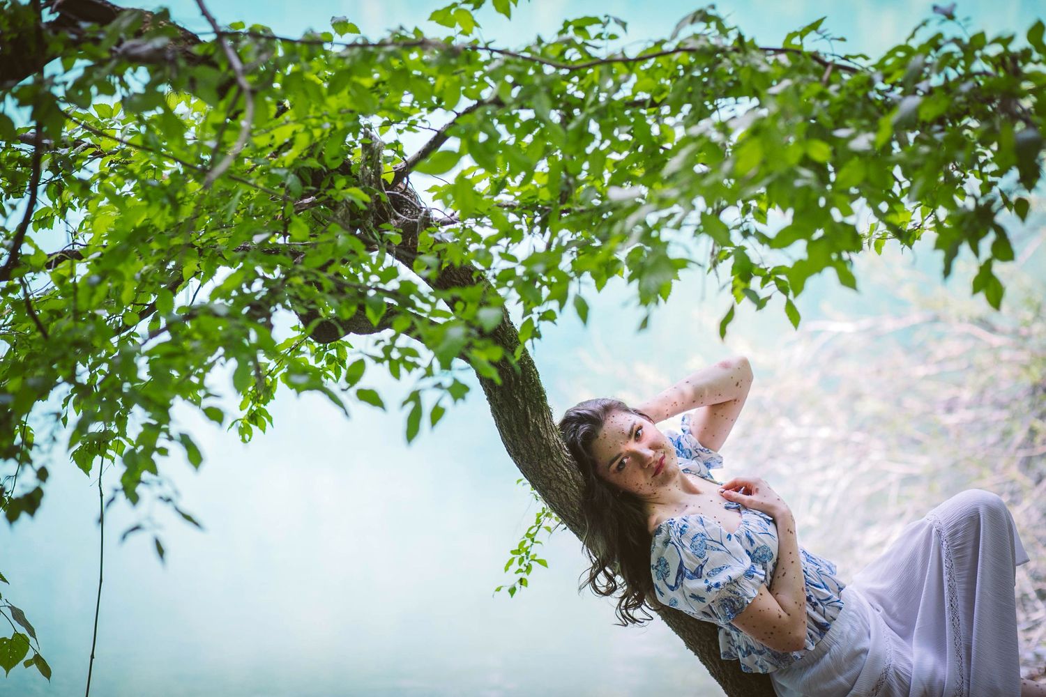 Artistic nature portrait under green tree branches beside tranquil water with ethereal lighting and flowing fabric.